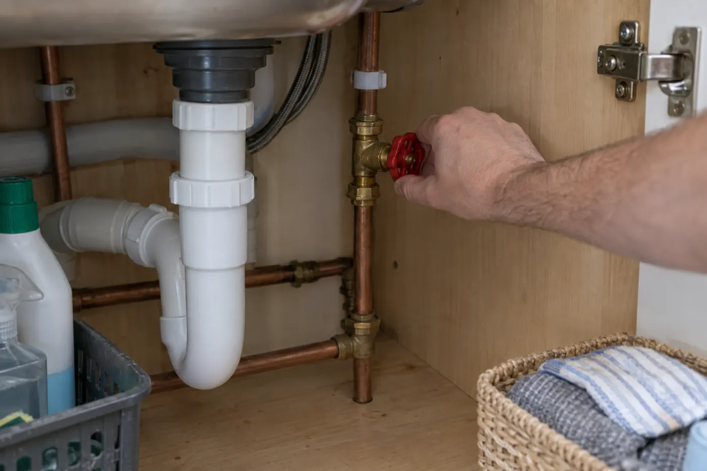 A hand turning the red handle of a stopcock under a kitchen sink
